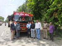 Seven men stand beside an orange transport truck on a sunny road, with lush green trees to the right; the truck has Hindi text on the windshield (likely a transport department name).