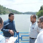 Four men stand on a blue railing by a lake, having a discussion; one holds papers and a phone.
