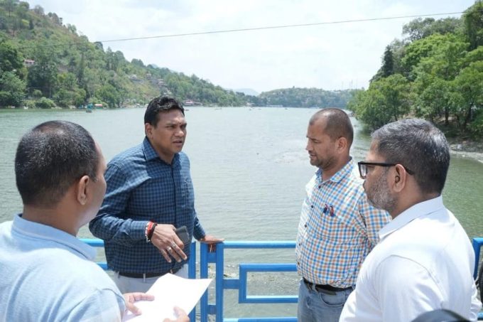 Four men stand on a blue railing by a lake, having a discussion; one holds papers and a phone.