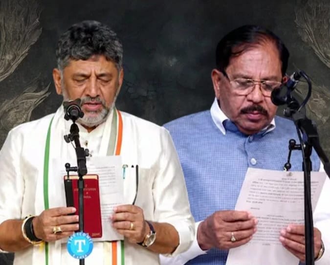 Two men stand at microphones reading from papers; the man on the left wears a white kurta with an Indian flag scarf and holds a red booklet, while the man on the right wears a light blue shirt and reads from a document.