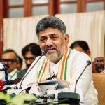 Senior man with gray beard speaking at a press conference, wearing white kurta and a tricolor scarf, microphones in front of him.