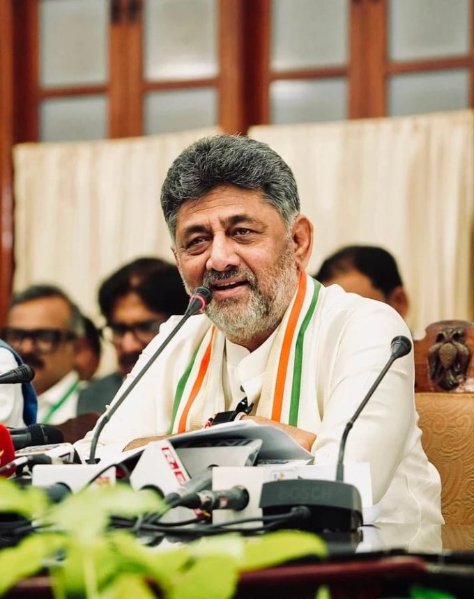 Senior man with gray beard speaking at a press conference, wearing white kurta and a tricolor scarf, microphones in front of him.