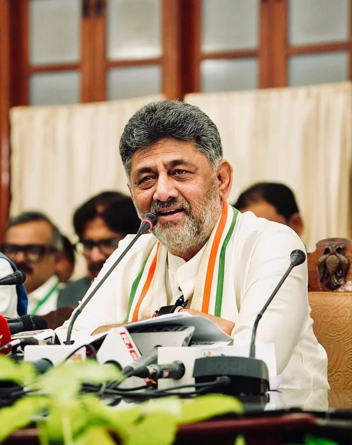 Senior man with gray beard speaking at a press conference, wearing white kurta and a tricolor scarf, microphones in front of him.