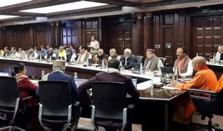 Long conference table with many people seated around, participating in a formal meeting in a wood-furnished room.]”,