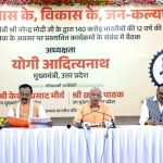 Panel of five men at a long table during a government event, with a large Hindi banner behind them celebrating 12 years of a program.