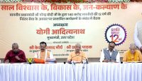 Panel of five men at a long table during a government event, with a large Hindi banner behind them celebrating 12 years of a program.