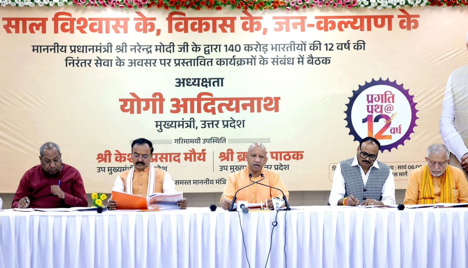 Panel of five men at a long table during a government event, with a large Hindi banner behind them celebrating 12 years of a program.