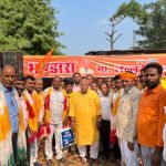 Group of Indian men in traditional attire posing at a political rally, orange banners in the background.