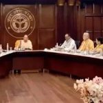 A formal government meeting in a wood-paneled room, with officials and Buddhist monks seated around a curved conference table; emblem on the back wall.
