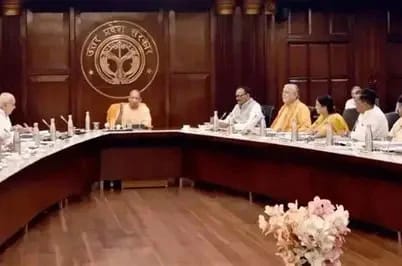 A formal government meeting in a wood-paneled room, with officials and Buddhist monks seated around a curved conference table; emblem on the back wall.
