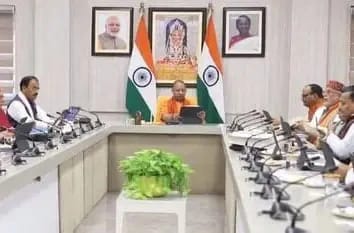 Formal government meeting in a conference room with Indian flags; central figure in orange robes chairs the panel, others seated along both sides.