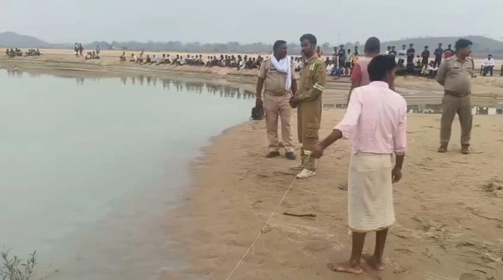 Man in a pink shirt and white wrap holds a rope along a sandy riverbank, with uniformed officers nearby and a line of spectators seated along the shore.