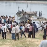 Large group of people gathered on a riverbank around a docked boat and several smaller boats on calm water.