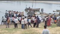 Large group of people gathered on a riverbank around a docked boat and several smaller boats on calm water.