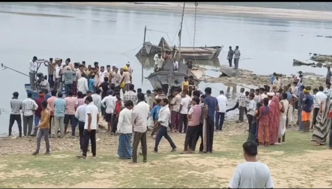 Large group of people gathered on a riverbank around a docked boat and several smaller boats on calm water.