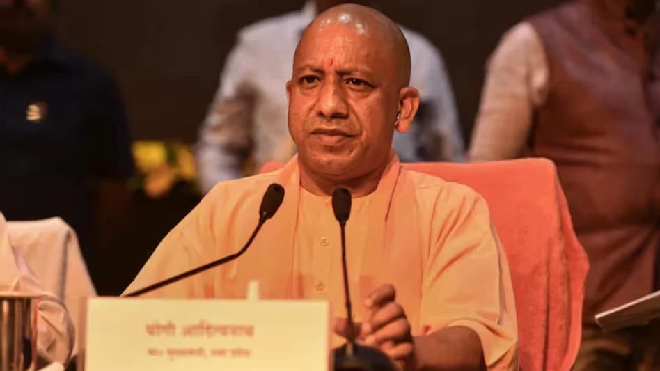 Man in peach traditional attire seated at a desk with two microphones, speaking at a formal event or panel discussion.