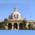 Front view of a grand beige building with a large central dome, arched entrance, and the Indian flag on top, palm trees in front.