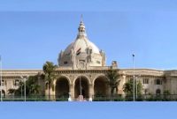 Front view of a grand beige building with a large central dome, arched entrance, and the Indian flag on top, palm trees in front.