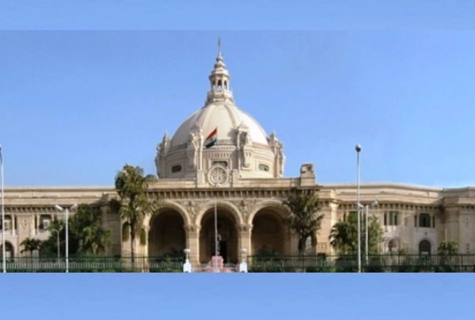 Front view of a grand beige building with a large central dome, arched entrance, and the Indian flag on top, palm trees in front.
