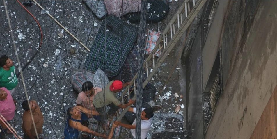 Group of people standing among concrete rubble and hanging wires, peering upward toward a damaged wall and ladder.