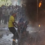 Man in a yellow shirt dodges flames as a protest scene burns nearby, with a crowd watching in the background.