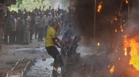 Man in a yellow shirt dodges flames as a protest scene burns nearby, with a crowd watching in the background.