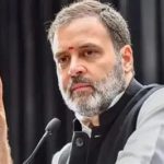 Bearded man in gray and black traditional attire speaks into a microphone, gesturing with his left hand.