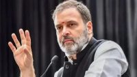 Bearded man in gray and black traditional attire speaks into a microphone, gesturing with his left hand.