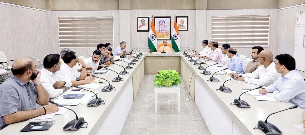 Formal government meeting in a conference room with a central speaker at the head of a U-shaped table, Indian flags and portraits behind him, attendees on both sides with documents and microphones.
