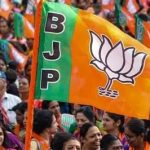 Crowd at a political rally waving a BJP flag with the lotus symbol and orange-green colors.”] ,