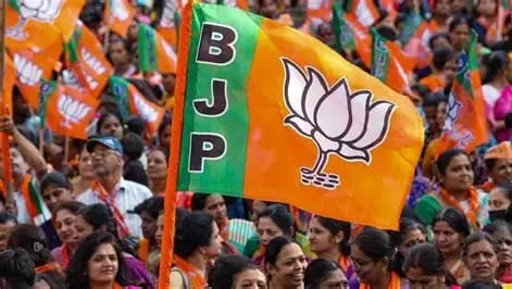 Crowd at a political rally waving a BJP flag with the lotus symbol and orange-green colors.”] ,