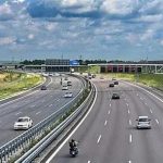 Multi-lane highway with cars and a motorcycle traveling under an overpass on a sunny day.