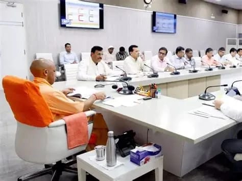 WhatsApp Image 2026-06-04 at 125737 AM - The Journalist News Monk in orange robes chairs at the head of a long conference table, addressing a group of men in white shirts in a modern meeting room, microphones on the table.