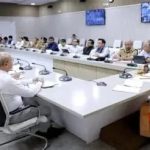 Panel of officials seated around a U-shaped conference table listening to a Buddhist monk in orange robes at the head of the room.