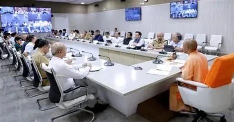Panel of officials seated around a U-shaped conference table listening to a Buddhist monk in orange robes at the head of the room.
