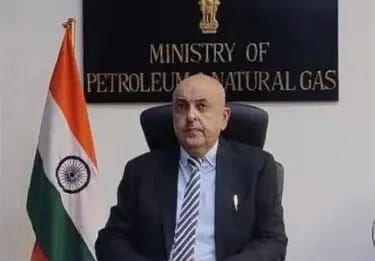 Bald man in a suit seated at a desk in front of a sign reading 'Ministry of Petroleum and Natural Gas' with the Indian flag beside him.
