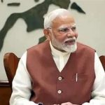 An older Indian man with white hair and beard wearing a brown sleeveless coat over a white kurta, seated at a desk in a formal setting, looking to the side.