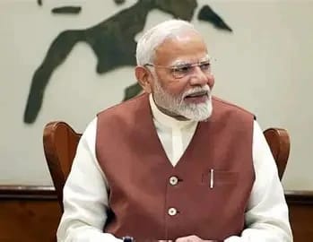 An older Indian man with white hair and beard wearing a brown sleeveless coat over a white kurta, seated at a desk in a formal setting, looking to the side.