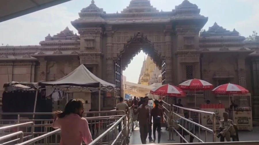 Ornate stone temple gateway with a carved arch, visitors walking along a ramp toward a distant spire, flanked by market stalls and red umbrellas on both sides.