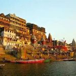 Colorful riverfront ghats with stairs and temple buildings, boats docked along the water, daytime scene.