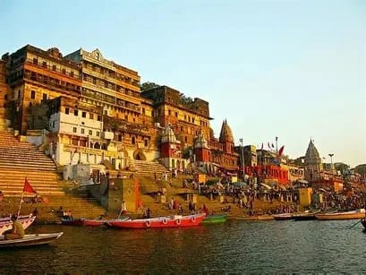 Colorful riverfront ghats with stairs and temple buildings, boats docked along the water, daytime scene.