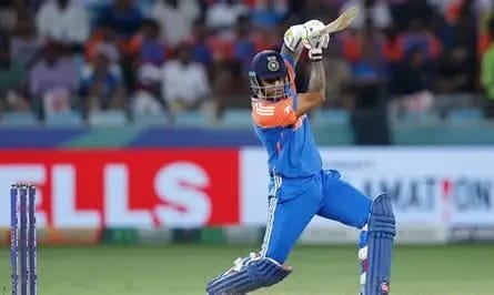 Cricketer in blue India jersey mid-swing against a cricket ball on a stadium field, bat raised for a shot.