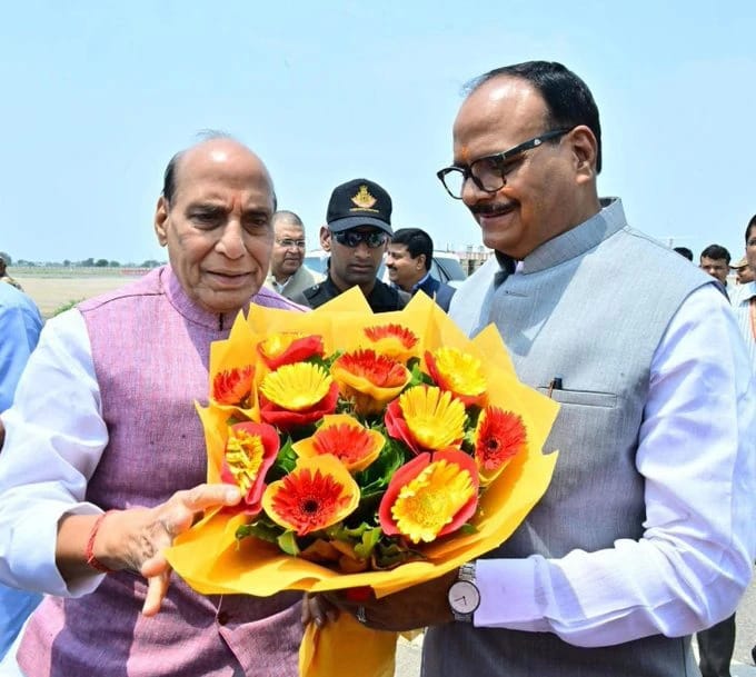 Two men exchange a bright bouquet of yellow and orange flowers outdoors, with onlookers in the background.