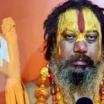 Bearded man with saffron-yellow face paint and a red vertical stripe, wearing orange bead necklaces, holding a ceremonial staff against an orange background