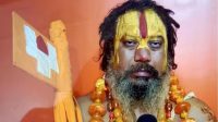 Bearded man with saffron-yellow face paint and a red vertical stripe, wearing orange bead necklaces, holding a ceremonial staff against an orange background