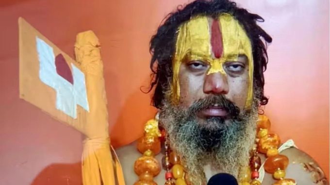 Bearded man with saffron-yellow face paint and a red vertical stripe, wearing orange bead necklaces, holding a ceremonial staff against an orange background