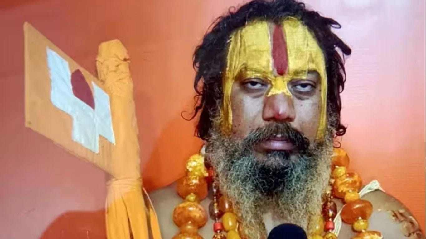 Bearded man with saffron-yellow face paint and a red vertical stripe, wearing orange bead necklaces, holding a ceremonial staff against an orange background