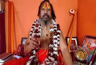 Bearded holy man in orange robes with flower garlands, seated in a bright orange room, pointing toward the camera.