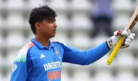 Cricketer in a blue India jersey raises his bat on the cricket field with a blurred stadium in the background.”