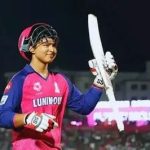 Cricketer in a pink jersey and helmet raises a bat in celebration on the cricket field.